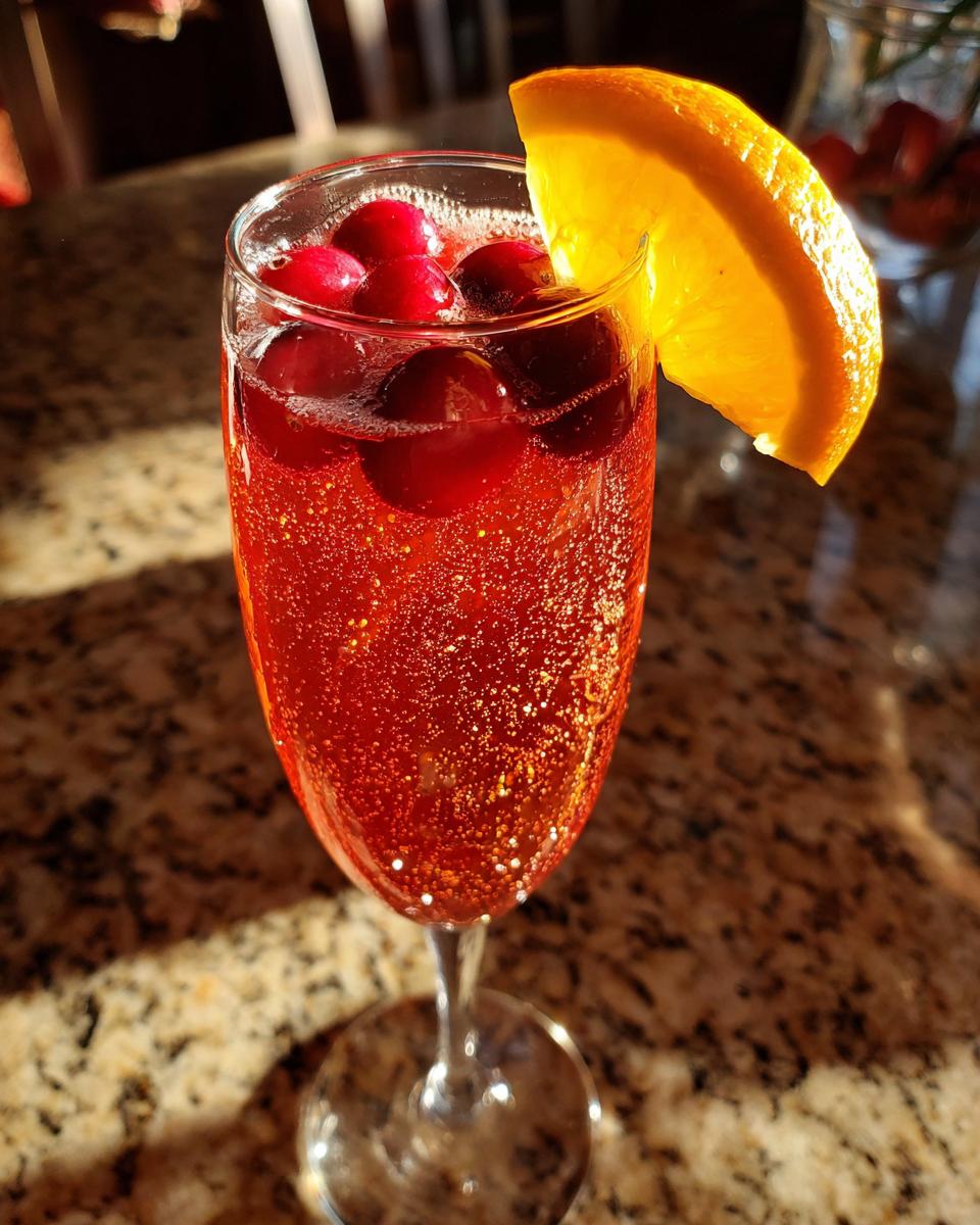 A sparkling Cranberry Mimosa cocktail in a champagne flute, garnished with fresh cranberries and an orange slice.